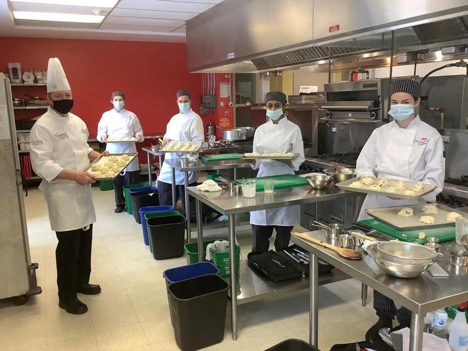 Culinary students are pictured in the culinary kitchen lab holding trays of meals they prepared. They are all wearing masks.