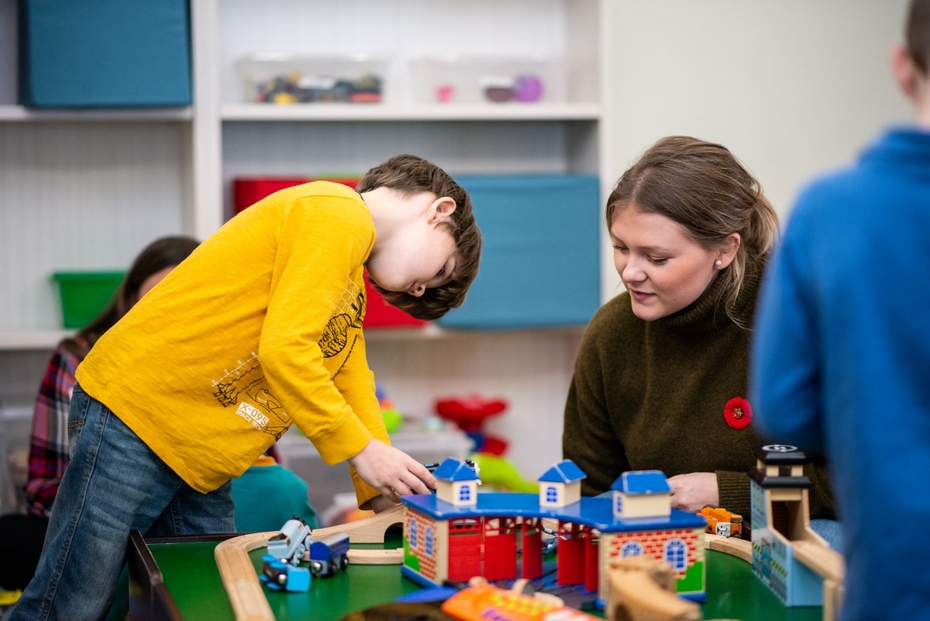A young boy plays with a train on a train table, while a woman watches.