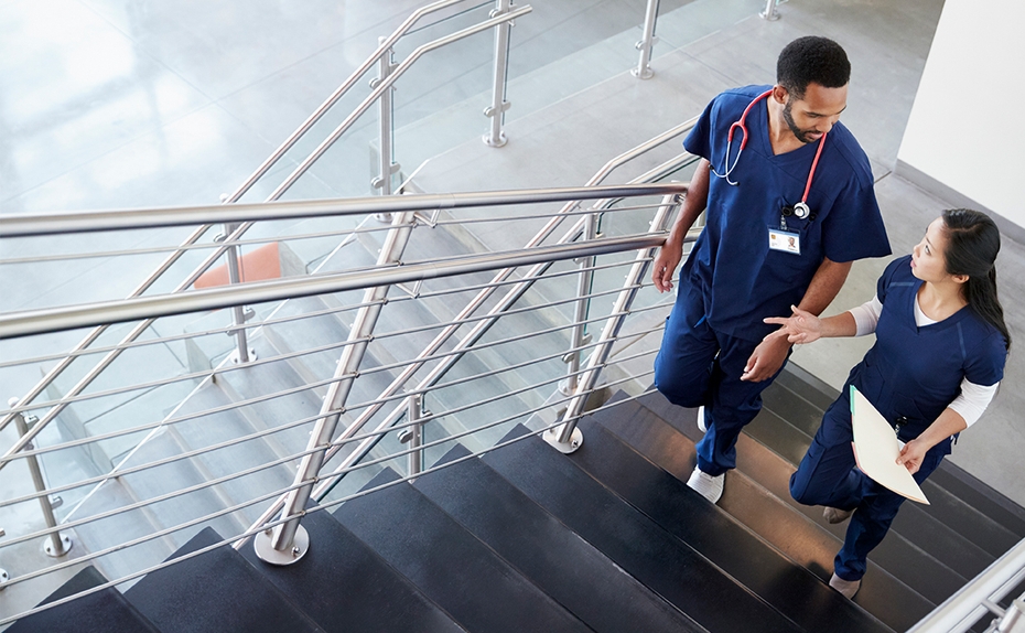 Two nurses in blue scrubs talk as they walk up stairs