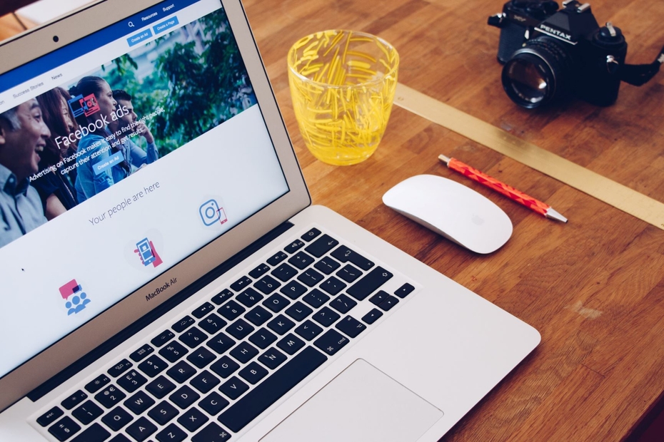 An open laptop screen shows Facebook ads manager. Beside the laptop are a camera, mouse, pen, and cup of water.