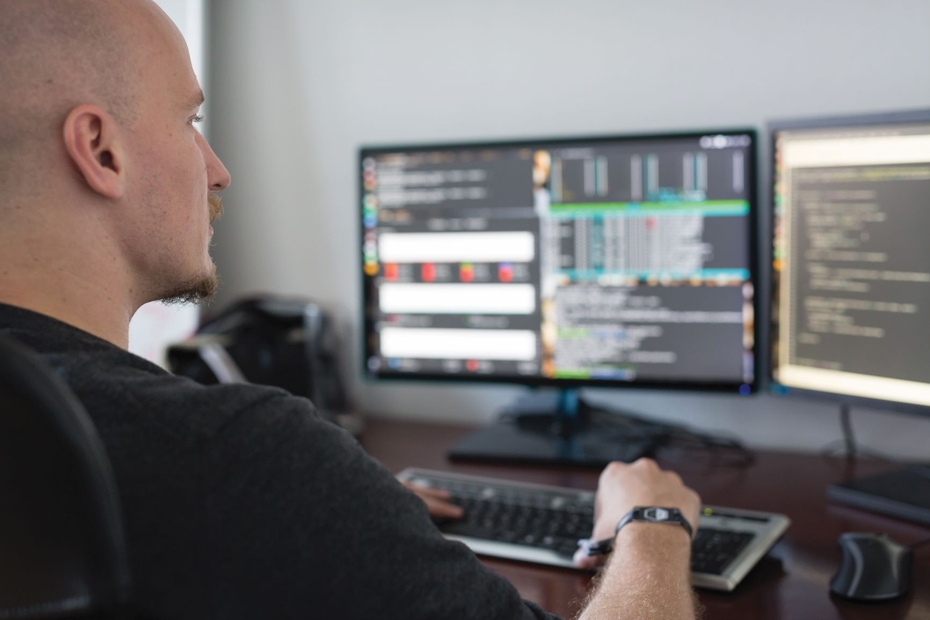 A man sits at a desk in front of dual computer screens, which display coding and programming.