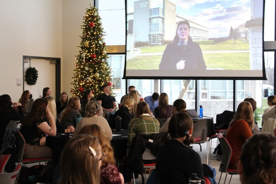 A student appears on the projector screen as tables of students and community partners watch the video in the SLC Event Venue