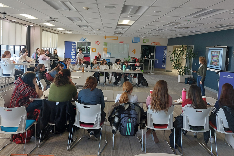 Students sit at tables around a classroom, listening to a speaker at the front of the room