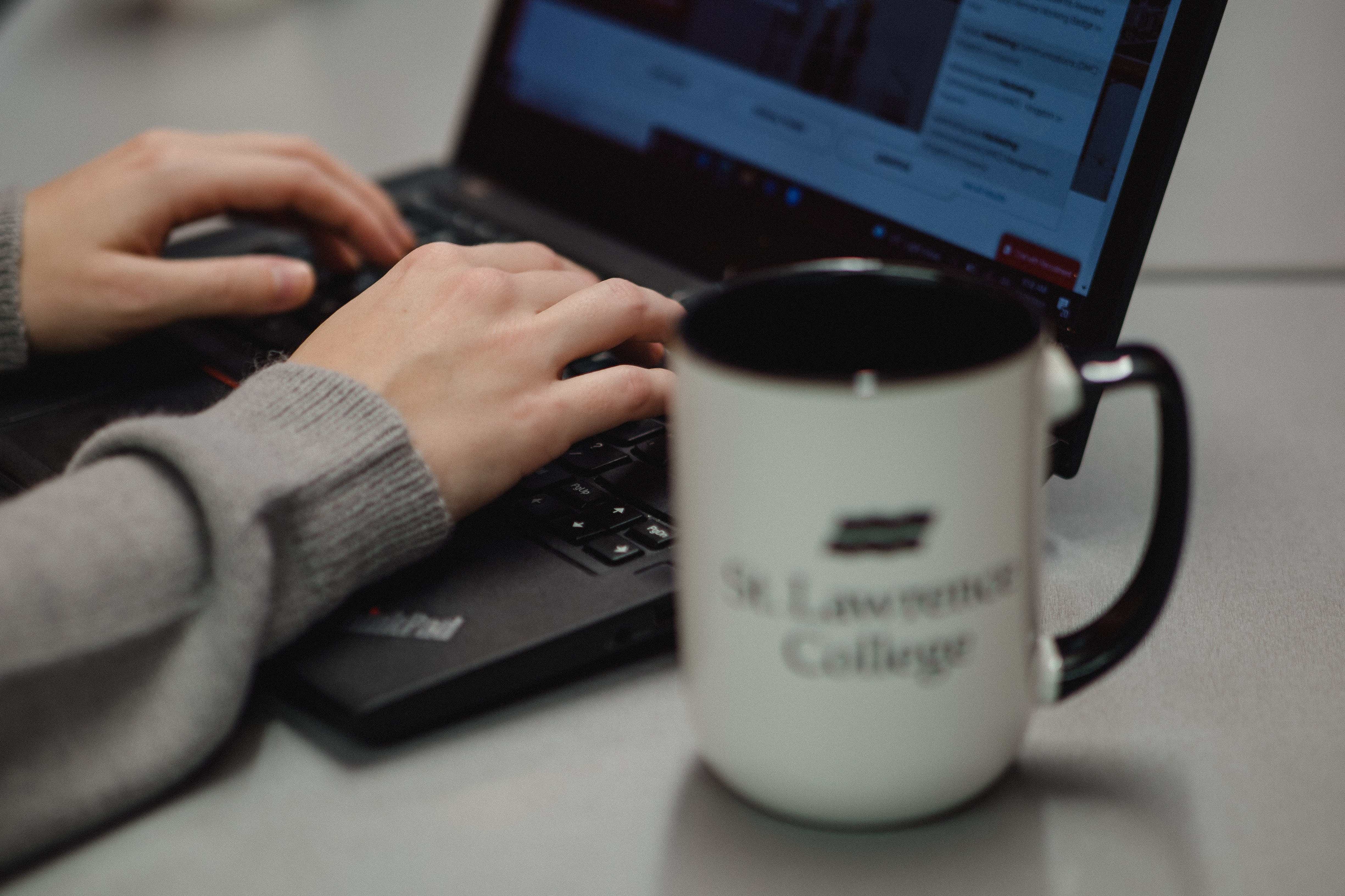 Hands typing on a laptop, St. Lawrence College mug on the side